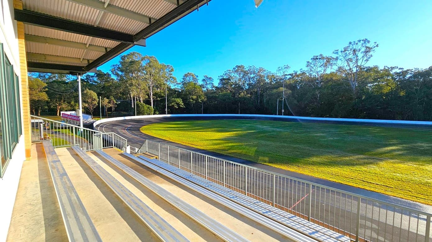 Metal bleachers on the left with a view of a cycling track curving around a grassy field. A white railing runs alongside the seating area. In the background, there are tall trees lining the perimeter of the track. A sign with "GOLDCOAST" is visible near the trees. The ceiling above the bleachers is made of corrugated metal with exposed beams. The floor is concrete.