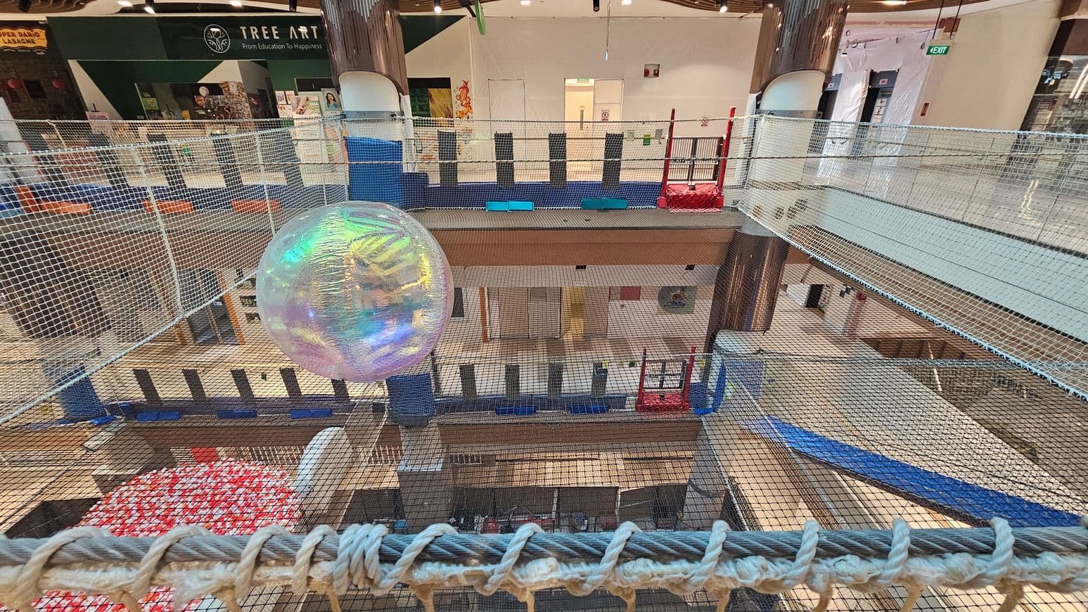A large, transparent, iridescent ball is suspended within a netted play area. The netting is extensive, covering multiple levels of a shopping centre. In the background, there is a shop with a sign reading "Tree Art" and various artworks displayed. The floor below is visible through the netting, showing a mix of open spaces and other structures. The area is brightly lit with overhead lights, and there are red and blue play structures on the right side.