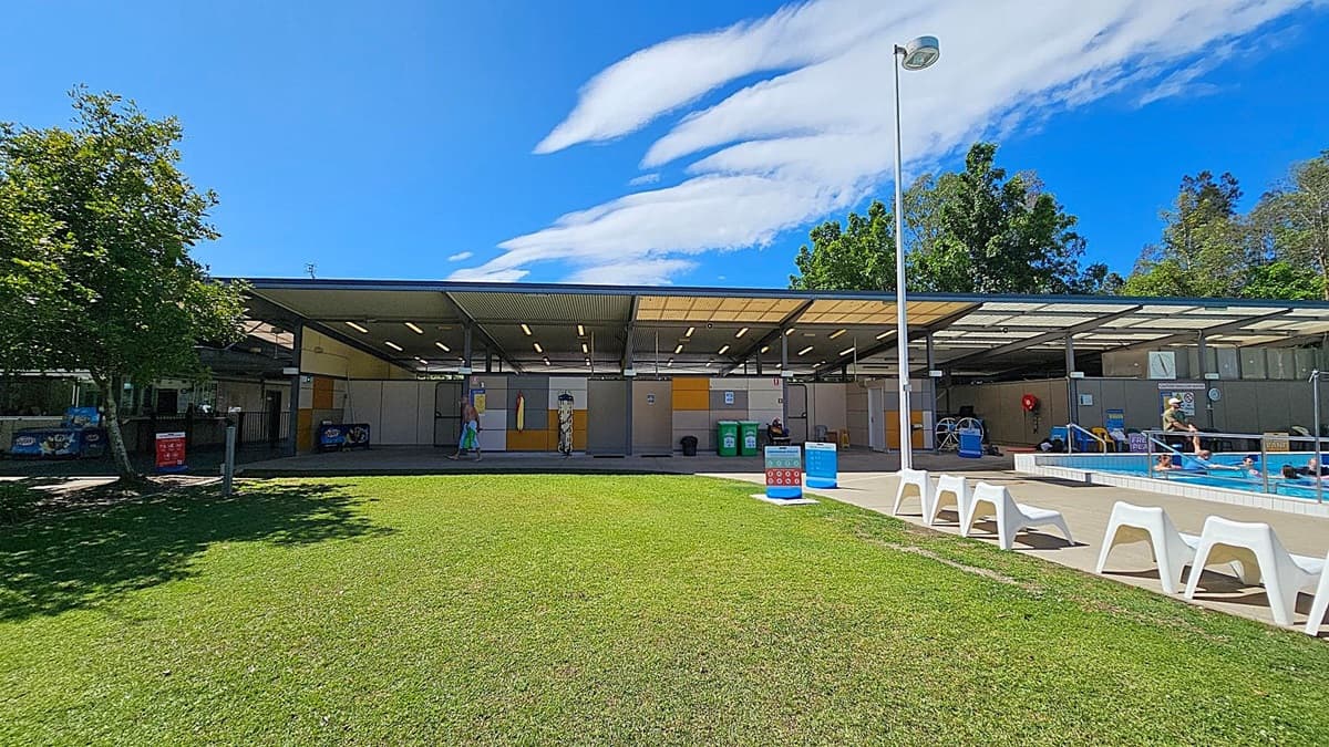 A grassy area in the foreground leads to a building with a corrugated metal roof in the background. On the left, there is a tree providing some shade. The building has open sections with people visible inside, and there are various signs and bins near the entrance. On the right, there is a swimming pool with people in it, and a row of white plastic lounge chairs along the poolside. A tall lamp post stands near the pool area. Trees are visible behind the building.