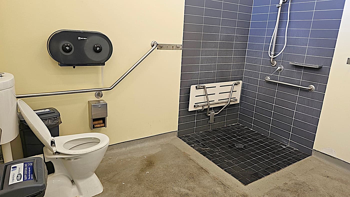 A bathroom with a toilet on the left, featuring a raised lid and a sanitary bin beside it. Above the toilet is a dual toilet paper dispenser and a grab bar. To the right is a shower area with dark grey tiles, a wall-mounted foldable shower seat, and multiple grab bars. The floor is a mix of concrete and tiled surfaces. The walls are painted light yellow.