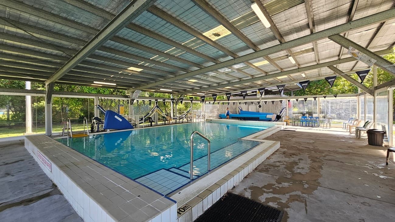 Indoor swimming pool with a tiled edge and clear water. The ceiling is made of metal beams with translucent panels, allowing natural light to filter through. On the left, there are large windows with a view of green trees and grass outside. On the right, there is pool equipment, including a blue pool cover roller. In the middle, a blue slide is visible at the far end of the pool. The floor around the pool is concrete with some wet patches. Black triangular flags are strung across the pool.
