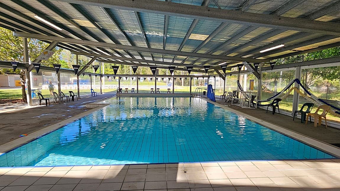 An indoor swimming pool with clear blue water, surrounded by a tiled floor. The ceiling is made of metal beams with translucent panels allowing natural light to filter through. On the left and right sides, there are plastic chairs placed along the pool's edge. The background features large windows with views of a grassy outdoor area and trees. Bunting with the word "Zoggs" is strung across the pool area. A lifeguard chair and some pool equipment are visible on the right side.