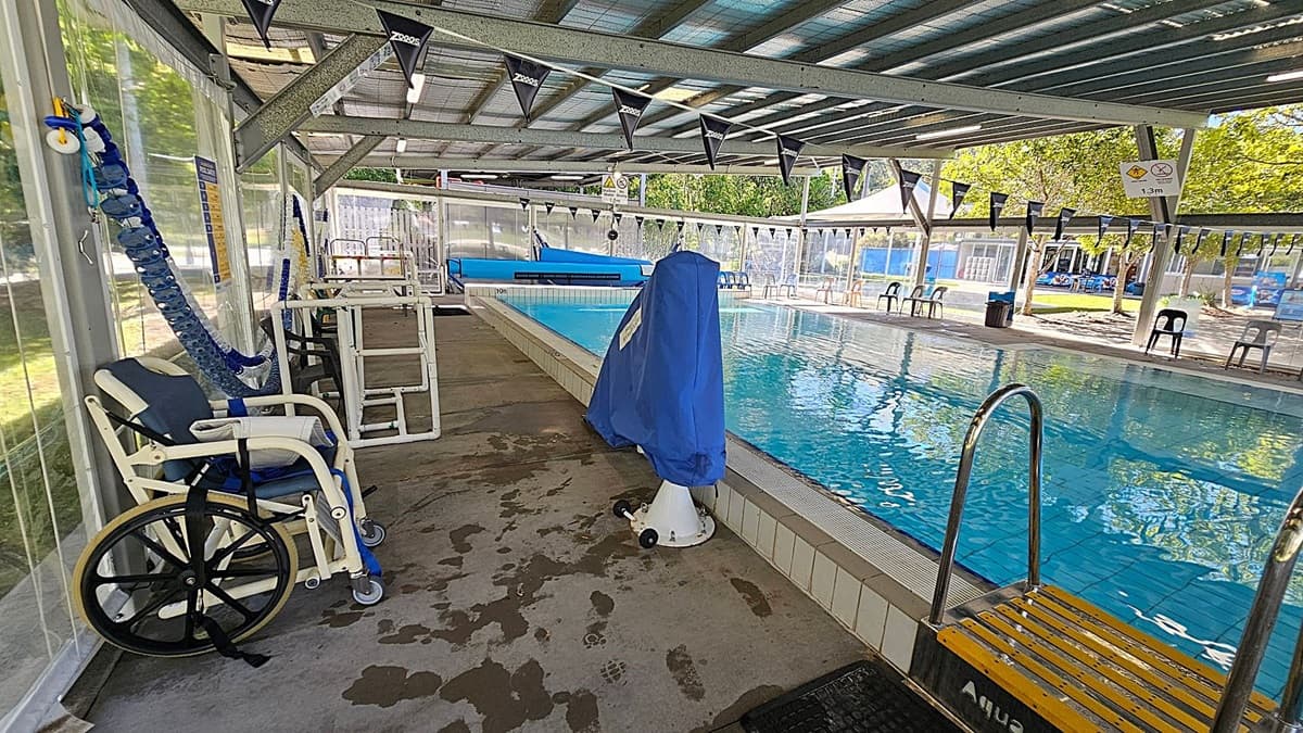 An indoor swimming pool area with a metal roof and transparent walls. On the left, there is a wheelchair and a blue pool lift. In the middle, the pool is visible with clear water, and a covered pool lift is positioned at the pool's edge. On the right, there are several plastic chairs lined up along the poolside. The floor is concrete with wet patches, and the ceiling has metal beams and lighting fixtures. Outside, trees and a playground area are visible through the transparent walls.