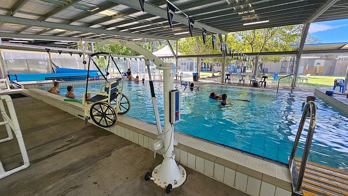 An indoor swimming pool with a metal roof and open sides. On the left, there is a pool lift with a wheelchair attached, and two people are sitting on the pool edge. In the middle, several people are swimming and engaging in water activities. On the right, there are metal handrails leading into the pool and a few plastic chairs and tables near the poolside. The floor is concrete, and the ceiling is made of metal sheets. Trees and a grassy area are visible outside.