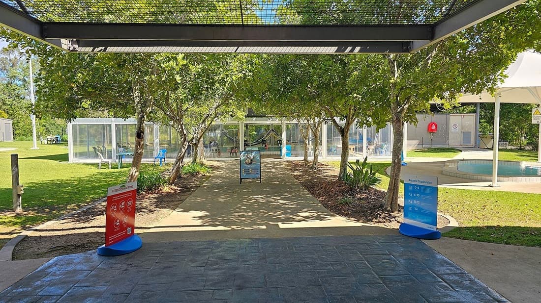 A paved pathway leads through a garden area with trees on both sides. In the foreground, there are two signs, one red and one blue, providing information. On the left, there is a grassy area with a transparent structure containing chairs. On the right, there is a small pool with a white canopy overhead. In the background, a glass building with people inside is visible. The floor is paved with stone tiles, and the ceiling is a metal grid structure.