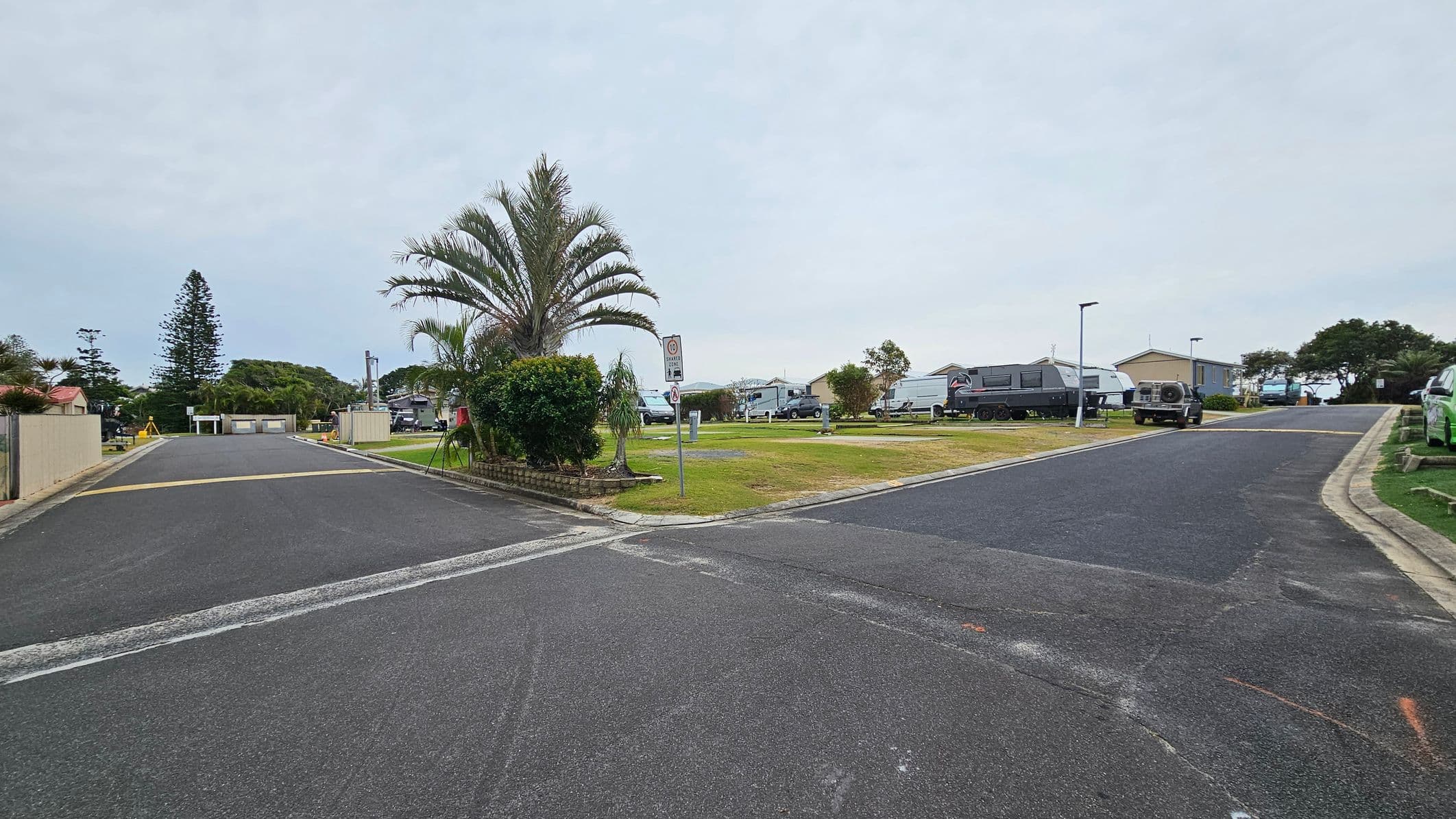 A paved road intersection with a palm tree and bushes on a grassy area in the centre. To the right, there are several caravans and a parked vehicle on a paved area. On the left, a road leads to a residential area with a tall conifer tree and more greenery. The roads are bordered by kerbs, and there are street lamps and a signpost near the centre. The sky is overcast.