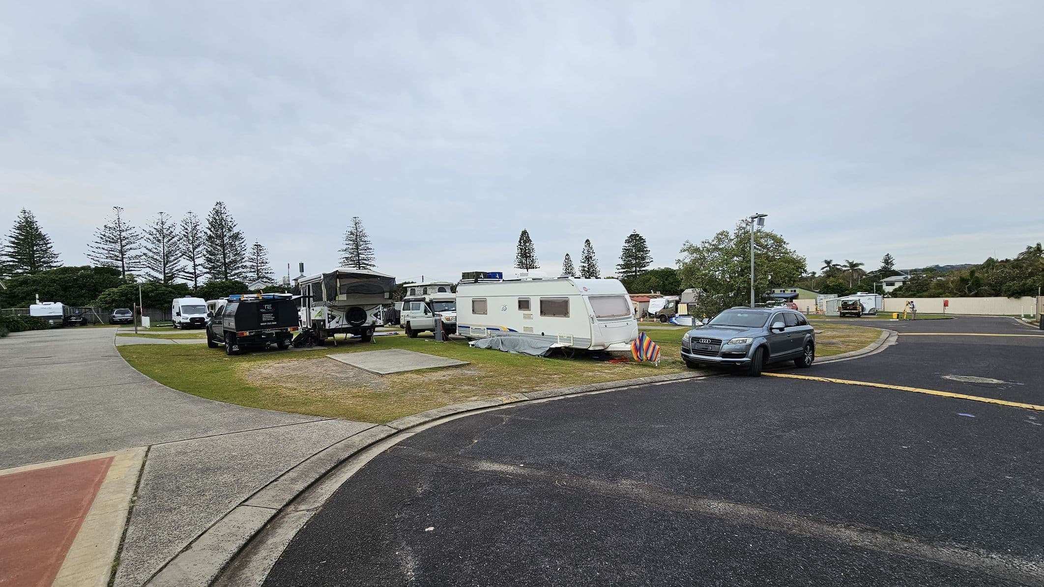 A caravan park with several vehicles and caravans parked on grassy areas. On the left, a black vehicle is attached to a pop-up camper, with a white van nearby. In the centre, a white caravan is parked with a colourful striped windbreak beside it. On the right, a grey SUV is parked on the edge of a paved road. In the background, tall trees and a few buildings are visible, with a light pole near the SUV. The ground is a mix of grass and asphalt.