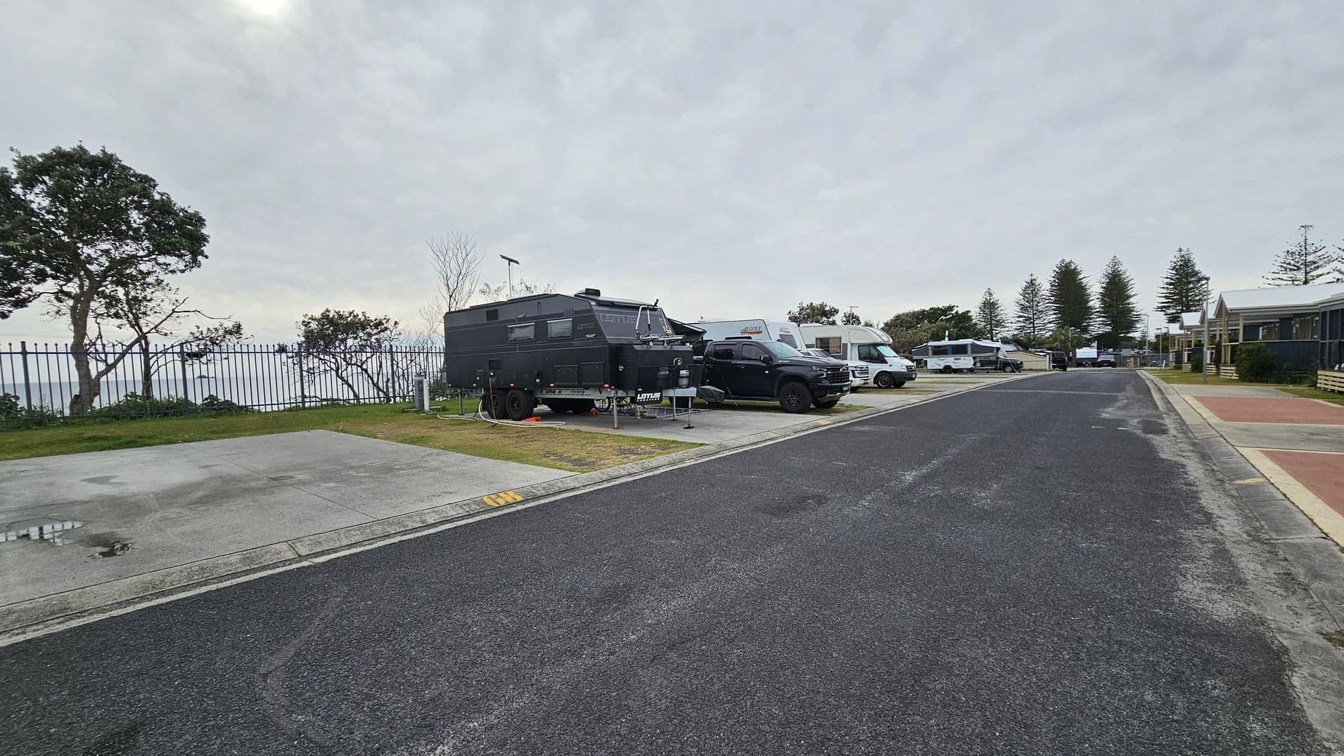 A paved road runs through a caravan park. On the left, a black caravan is parked next to a black vehicle, with more caravans lined up behind. A metal fence and trees are visible in the background. On the right, there are more caravans and a building with a red-tiled area in front. The ground is a mix of asphalt and grass, and the sky is overcast.