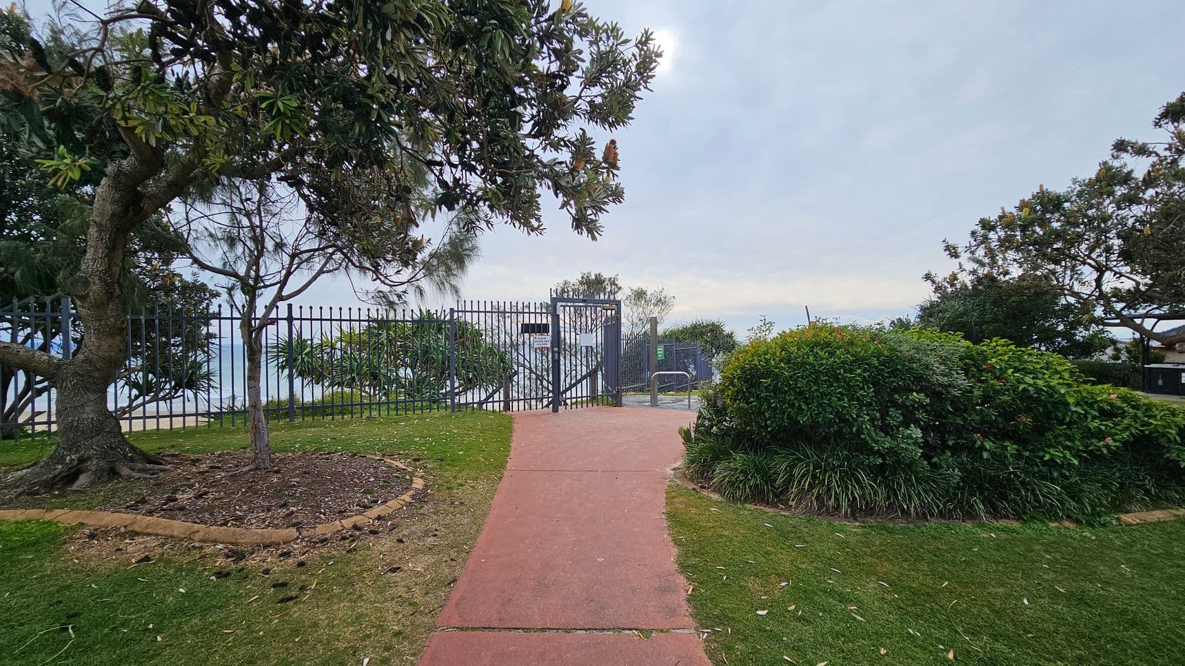A pathway made of reddish concrete leads to a metal gate in the centre, surrounded by a tall fence. On the left, a large tree with a thick trunk and overhanging branches is bordered by a circular patch of soil. On the right, dense green bushes and plants line the path. In the background, more trees and foliage are visible beyond the fence, with a hint of water or sky beyond. The ground is covered with grass, and the area appears well-maintained.