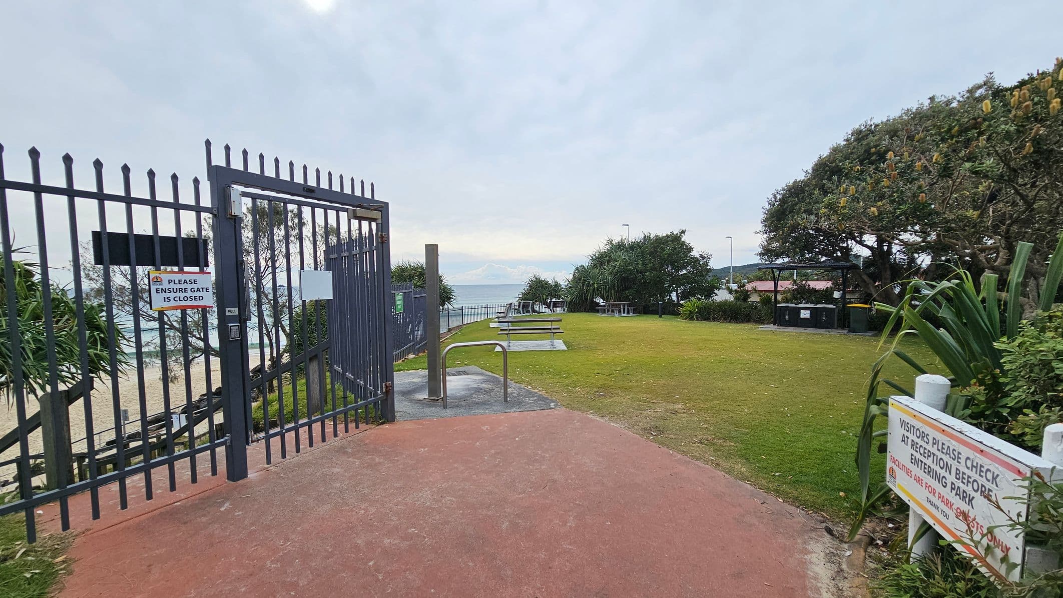 A metal gate on the left with a sign reading "PLEASE ENSURE GATE IS CLOSED" opens to a path leading to a beach. The path is bordered by grass and a few picnic tables. On the right, a sign instructs visitors to check in at reception before entering the park. Trees and bushes are scattered around, with a view of the ocean in the background. The ground is a mix of red and grey pavement, and the area is open and grassy.