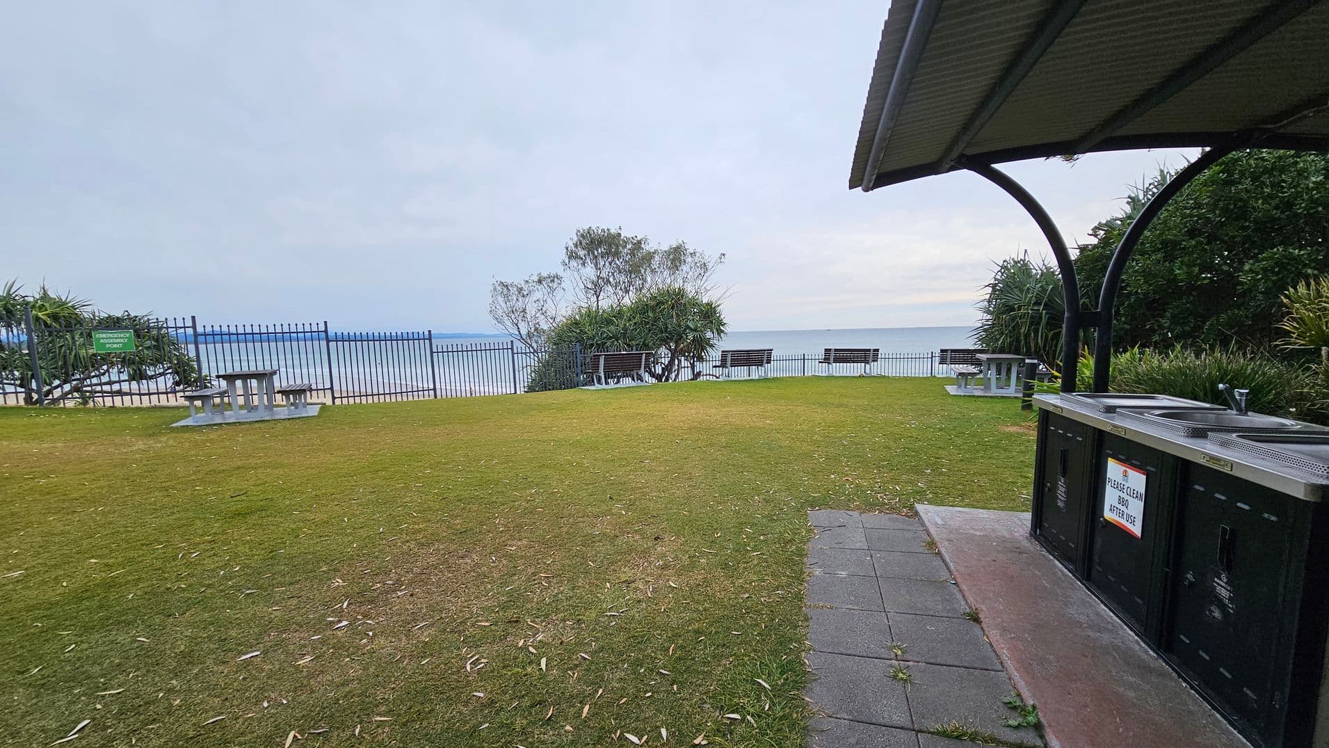 A grassy area with a few picnic tables and benches facing the ocean. On the left, a metal fence runs along the edge of the grass, with a sign that reads "Observation Point." In the foreground on the right, there is a covered barbecue area with a sign asking to clean after use. The background features the ocean and some trees and shrubs. The ground is a mix of grass and a paved path near the barbecue.