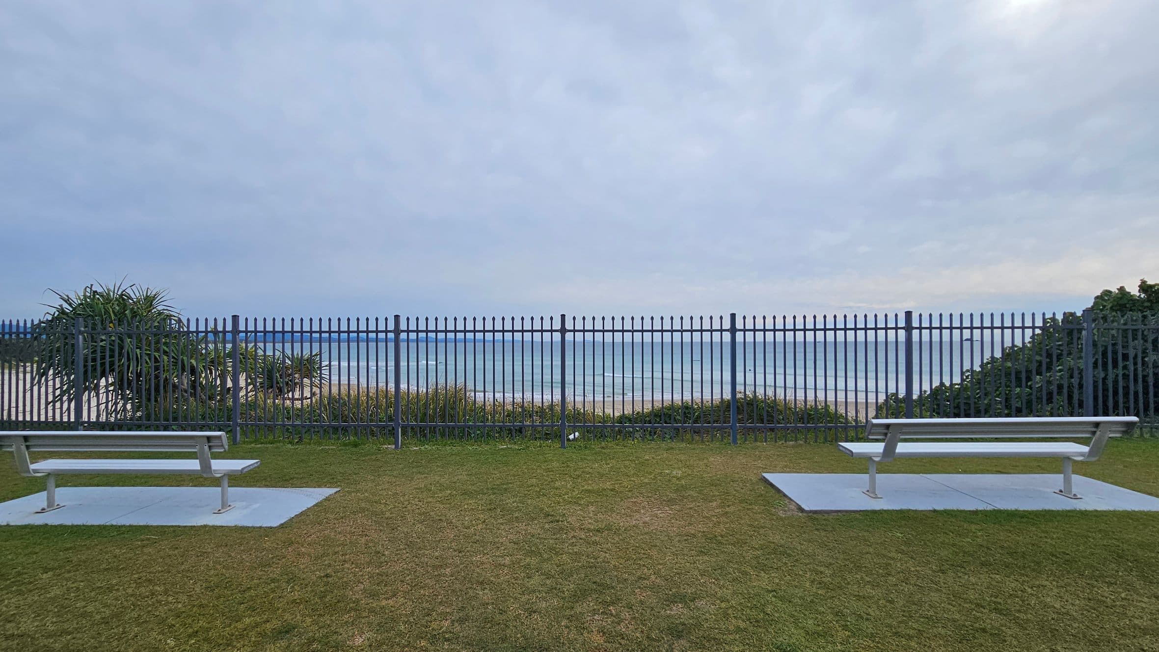 Two metal benches are positioned on concrete slabs in a grassy area. A tall metal fence runs horizontally across the image, separating the benches from the view beyond. In the background, the ocean is visible, with waves gently rolling in. On the left, there are some leafy plants, while on the right, more greenery is present. The sky is overcast, casting a muted light over the scene.