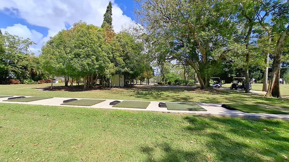On the left, a well-maintained grassy area extends towards a line of trees, providing a natural boundary. In the centre, a gravel pathway leads into the distance, flanked by more greenery. On the right, a fenced area contains golf mats and a small practice area, with a few trees nearby. The ground is primarily gravel with patches of grass.