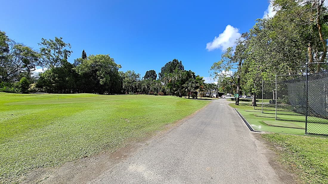 On the left, a large expanse of well-maintained grass stretches towards a line of trees, providing a natural backdrop. In the centre, a gravel pathway runs straight ahead, leading into the distance. On the right, a fenced area contains golf mats and a small practice space, with a few trees nearby. The ground is primarily gravel with patches of grass.