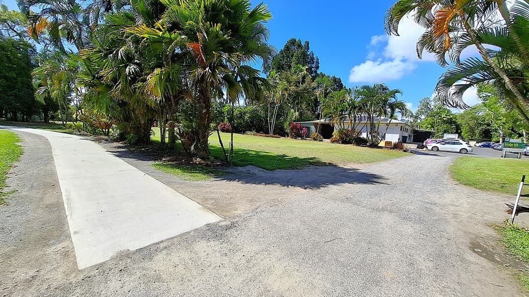 On the left, a concrete pathway curves gently, leading into a grassy area bordered by palm trees and shrubs. In the centre, a flat expanse of lawn is visible, with a small building partially obscured by greenery. On the right, a gravel road branches off, with a parked car and a sign indicating "Hole 10" nearby. The ground is a mix of concrete, gravel, and grass.
