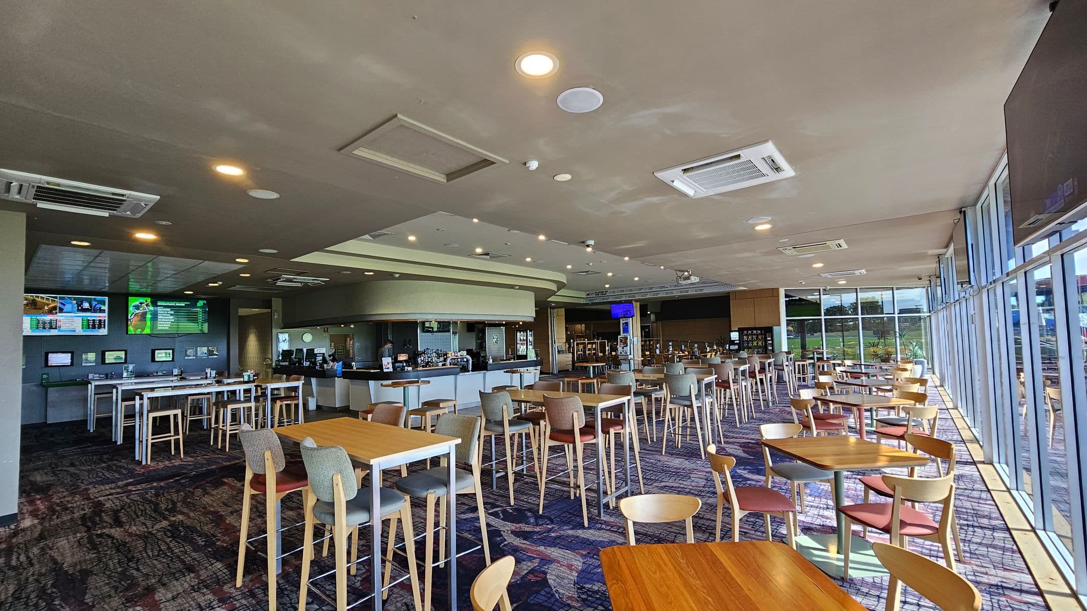 A spacious, modern dining area with a mix of high and low wooden tables and chairs. On the left, there are several screens mounted on the wall above a long counter. The centre features a bar area with stools. The right side has large windows allowing natural light to flood in, with views of greenery outside. The floor is carpeted with a patterned design, and the ceiling is fitted with recessed lighting and air conditioning units.
