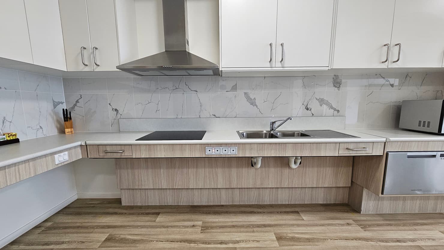 Inside the accommodation is a kitchen area furnished with wooden cabinets, a kitchen sink, a microwave, and a small fridge, complete with a cooktop and kitchen hood. Kitchen accessories are neatly placed in the corner. The floor is timber, and the ceiling is white with LED lights.