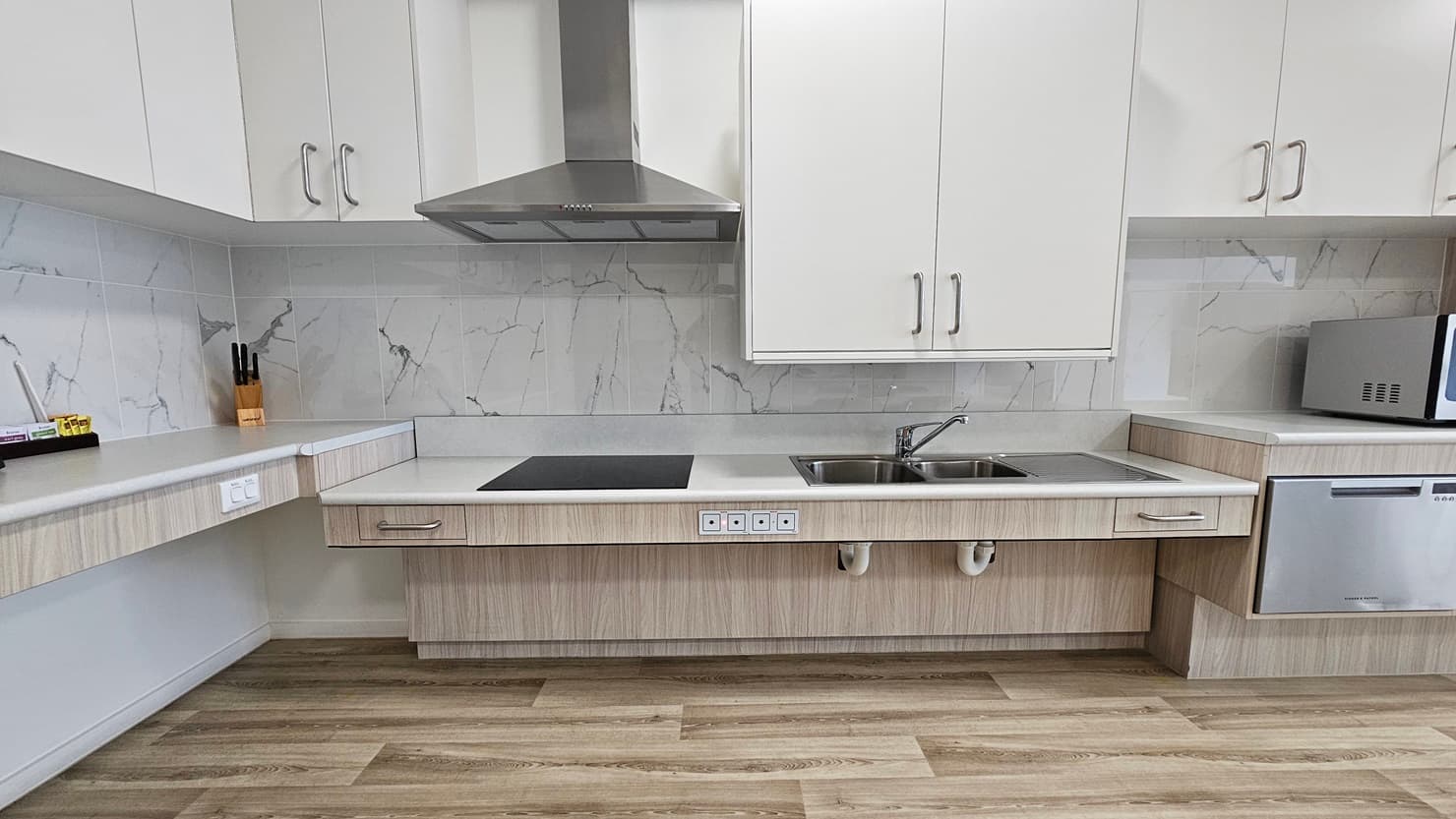 Inside the accommodation, find a kitchen area furnished with wooden cabinets, a kitchen sink, a microwave, and a small fridge, complete with a cooktop and kitchen hood which has been lowered for easier access. Kitchen accessories are neatly placed in the corner. The floor is timber, and the ceiling is white with LED lights.