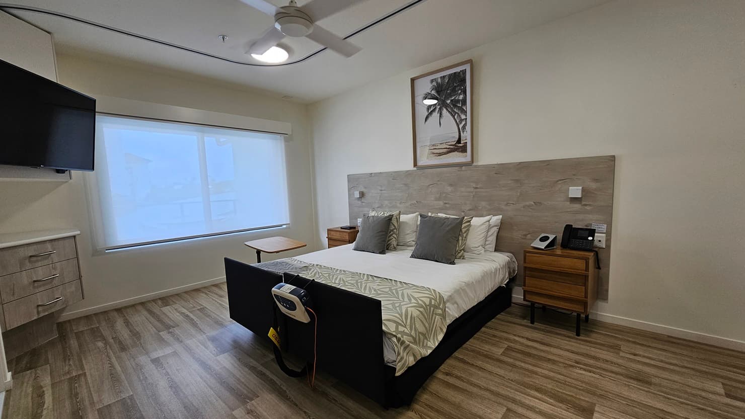 In the middle is a double bed with a wooden headrest. Wooden cabinets flank both sides, one holding a telephone. A painting hangs above the headrest. To the left is a large window with a flatscreen TV mounted on the wall. Below it is another wooden cabinet. The floor is timber, and the wall and ceiling are white with an LED light and a ceiling fan.