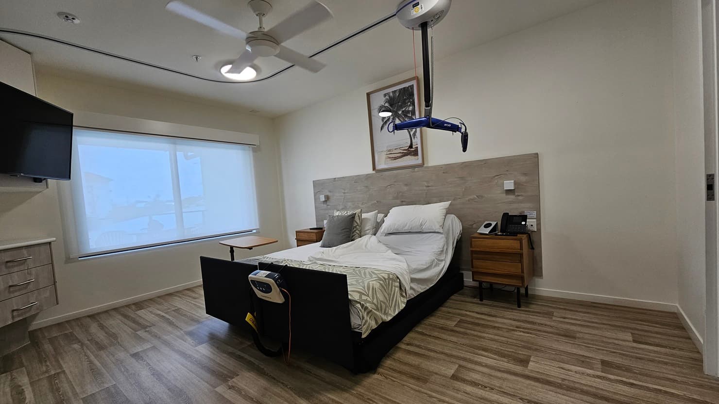 In the middle is a double bed with a wooden bedhead. Wooden cabinets flank both sides, one holding a telephone. A painting hangs above the headrest. A hoist hangs from the ceiling, and a monitor is attached to the bed. To the left is a large window with a flatscreen TV mounted on the wall. Below it is another wooden cabinet. The floor is timber, and the wall and ceiling are white with an LED light and a ceiling fan.