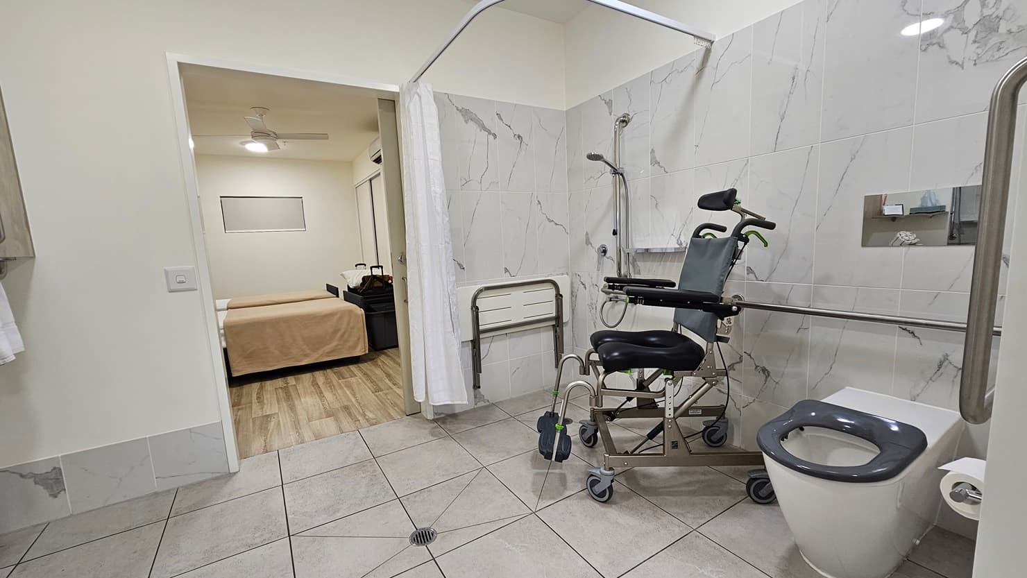 In the ensuite bathroom, an aquatic wheelchair sits in the corner near the shower. A foldable bench chair is mounted to the wall, with a curtain hanging from above. To the right is a toilet bowl equipped with handrails and a tissue dispenser, with a mirror above it. The walls have white tiles, and the ceiling is also white. On the left is the bedroom area.