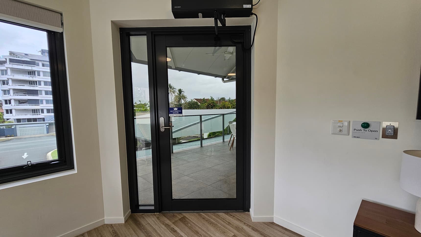 Access the accommodation through an automatic glass door with a "Push To Open" button on the right. A TV stand and a white lampshade stand on the right. To the left is a window offering a view of the outside. The floor is made of timber, and the walls are all white.