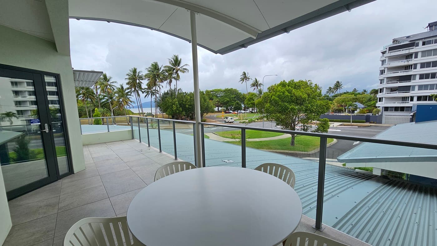 The balcony features a round table and four movable plastic chairs in the middle. The roof extends for additional shade, with glass railings securing the area. To the left is a glass door, while tall palm trees line the left side along with other trees. To the right is the road and a large building.