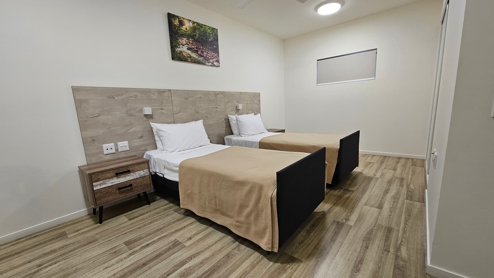In the middle are two single beds placed side by side, each with a bedside table and a wooden headrest. A painting hangs above. The walls and ceilings are all white with LED lights. The floor is made of timber.