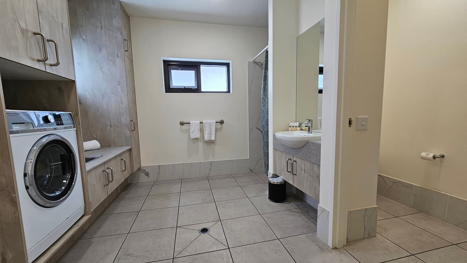 The laundry area has a washing machine on the left and a sink beside it. Two small windows are in the middle, with a hand grab and two white towels below. On the right is a wash basin, toilet bowl, and shower area.  A rubbish bin is placed under the wash basin. The floor has grey tiles, and the ceiling and walls are all white.