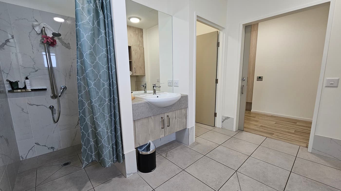 On the right is a shower with a green shower curtain and a shampoo holder. Beside it is a washbasin with a large mirror. Underneath is a black rubbish bin. Adjacent is an inward-opening door. The floor has grey tiles, and the ceiling and walls are all white with a black accent. To the right is a door and a timber floor is visible.