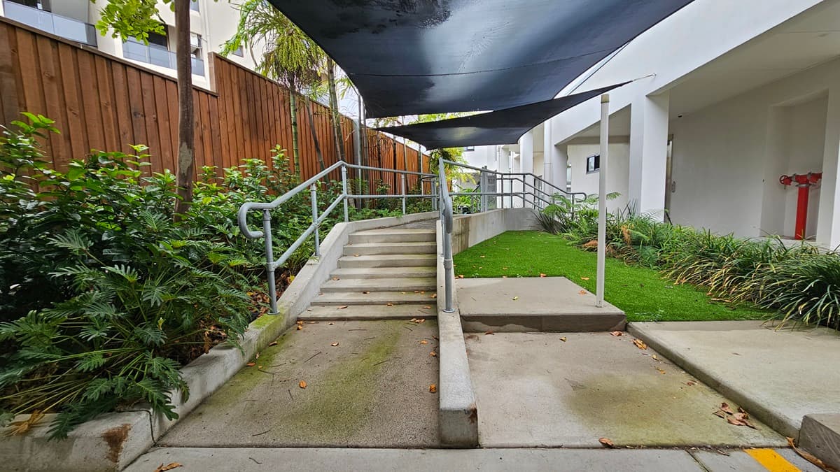 In the shelter of a corrugated roof lies accessible parking, marked by a blue wheelchair sign and yellow diagonal lines. Opposite are three vehicles of different models parked. On the right stands a white building, enclosed by a wooden fence. The area is surrounded by trees and ornamental plants.