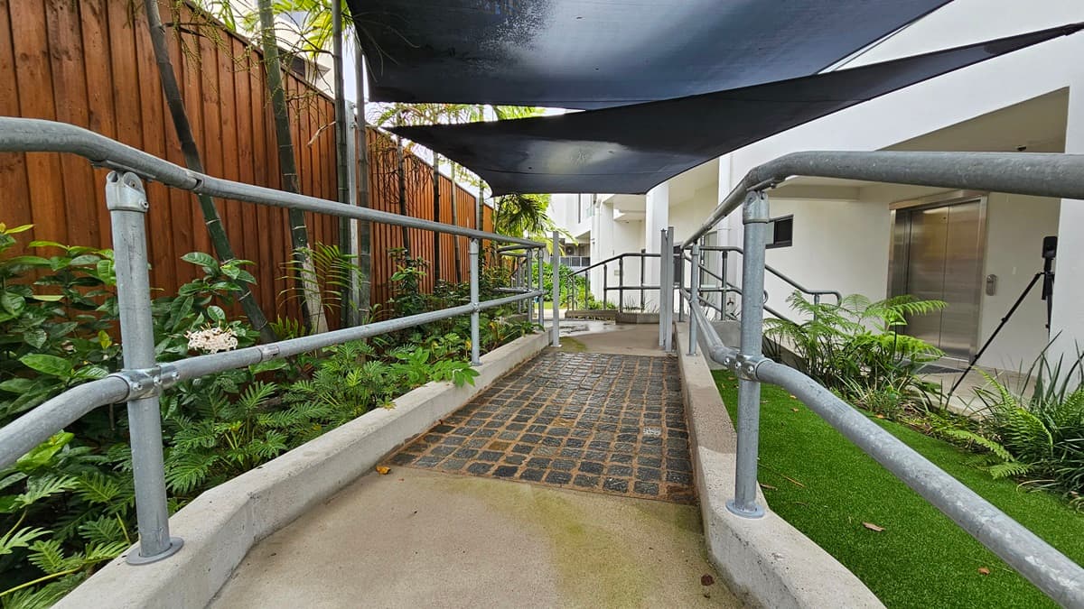 In the middle is a concrete ramp made of black tiles with metal handrails. Landscape plants and manicured grass line both sides. Overhead, a black shade cloth offers cover. The ramp descends to stairs on the right, with a lift nearby. On the left, a wooden plank fence encloses the area.