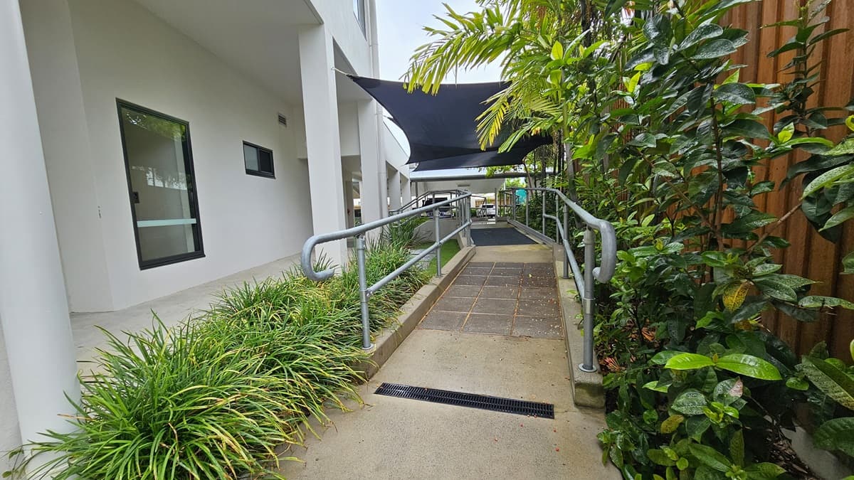 In the centre is a concrete ramp with grey tile flooring and metal handrails. Landscaped plants and manicured grass flank both sides. Above, a black shade cloth provides cover. To the right, a wooden plank fence encloses the area. On the left stands the facility building with two glass windows.