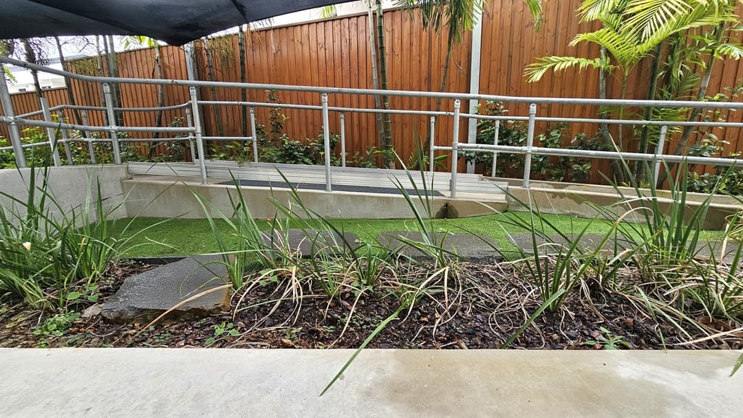 A concrete ramp with metal handrails runs between landscaped plants and manicured grass. A black shade cloth provides overhead cover, while a wooden plank fence encloses the area.