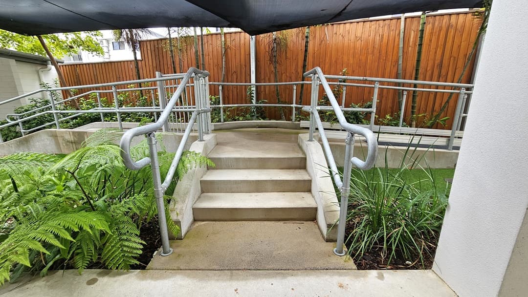 A concrete staircase with metal handrails is flanked by landscaped plants and manicured grass. Overhead, a black shade cloth provides cover, while a wooden plank fence encloses the area.