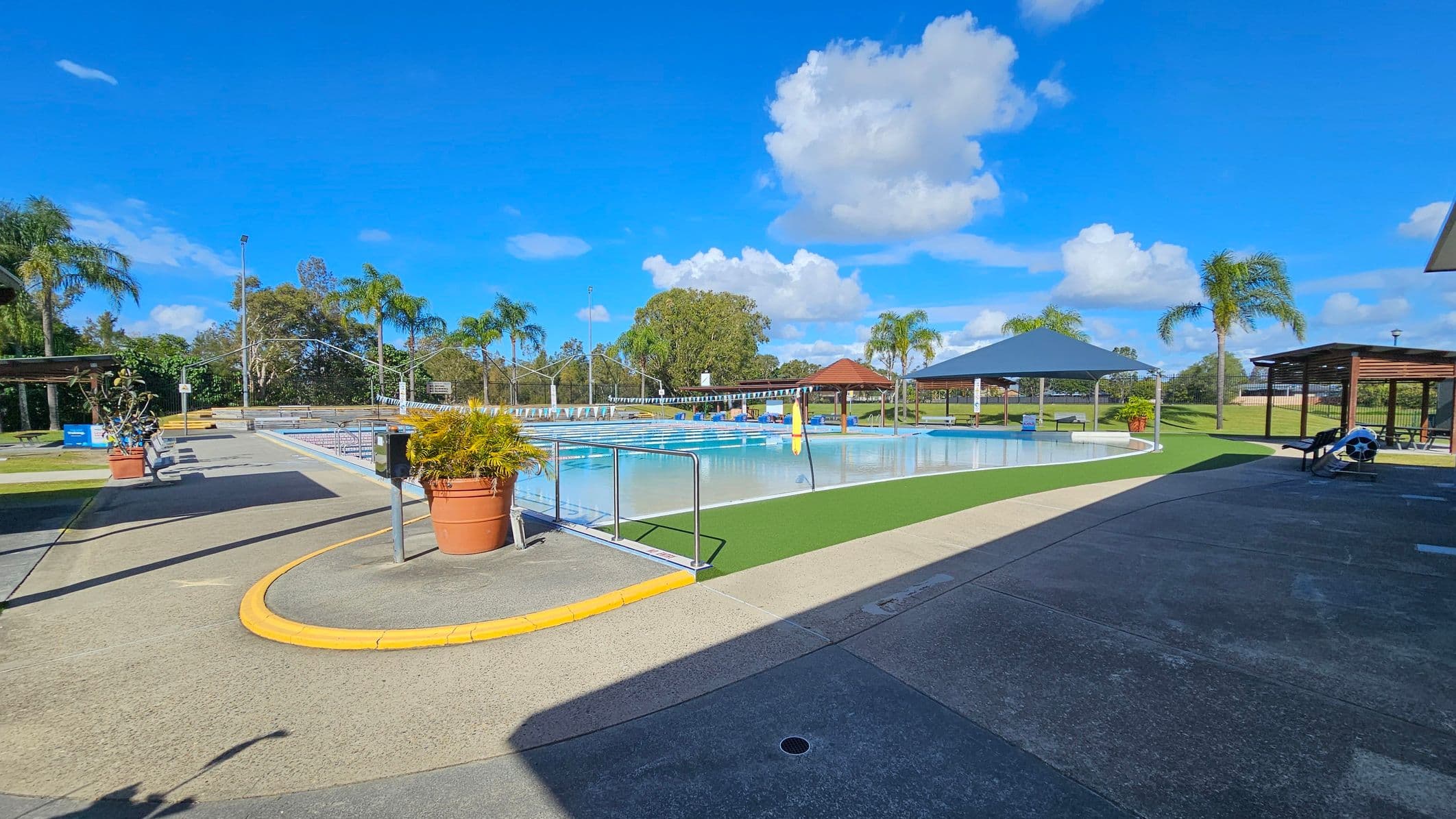 An outdoor swimming pool area with a clear blue sky. On the left, there are potted plants and a pathway leading to the pool. In the centre, the pool is surrounded by a green artificial turf. To the right, there are shaded seating areas with wooden structures and a large blue canopy. Palm trees are scattered throughout the background, and the ground is a mix of concrete and green turf.