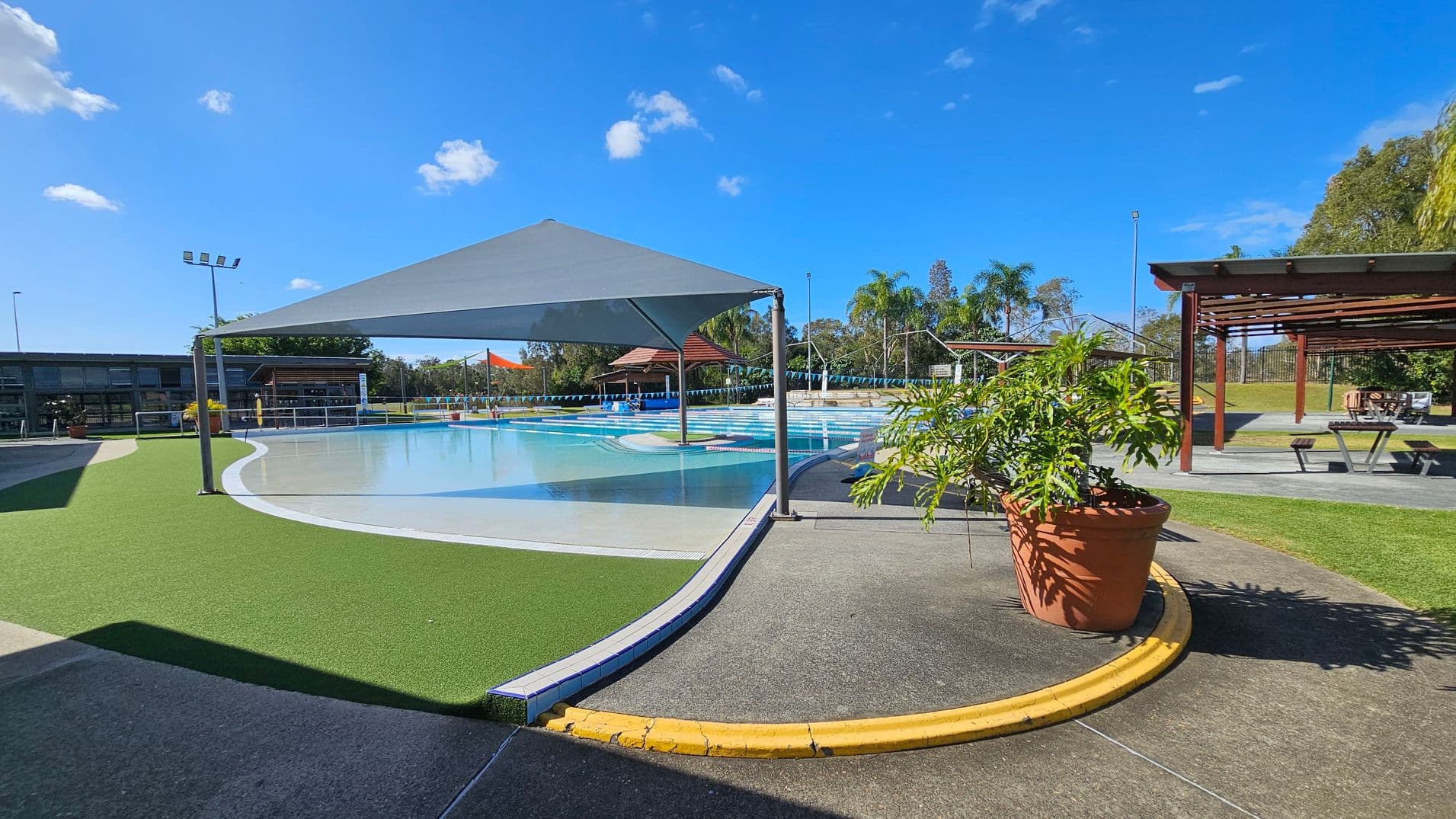 A swimming pool area with a shade structure over part of the pool. The pool is surrounded by artificial grass and concrete paths. On the right, there is a large potted plant and a picnic area with tables under wooden pergolas. In the background, there are more shaded areas, a building with large windows, and some palm trees. The sky is clear with a few clouds.