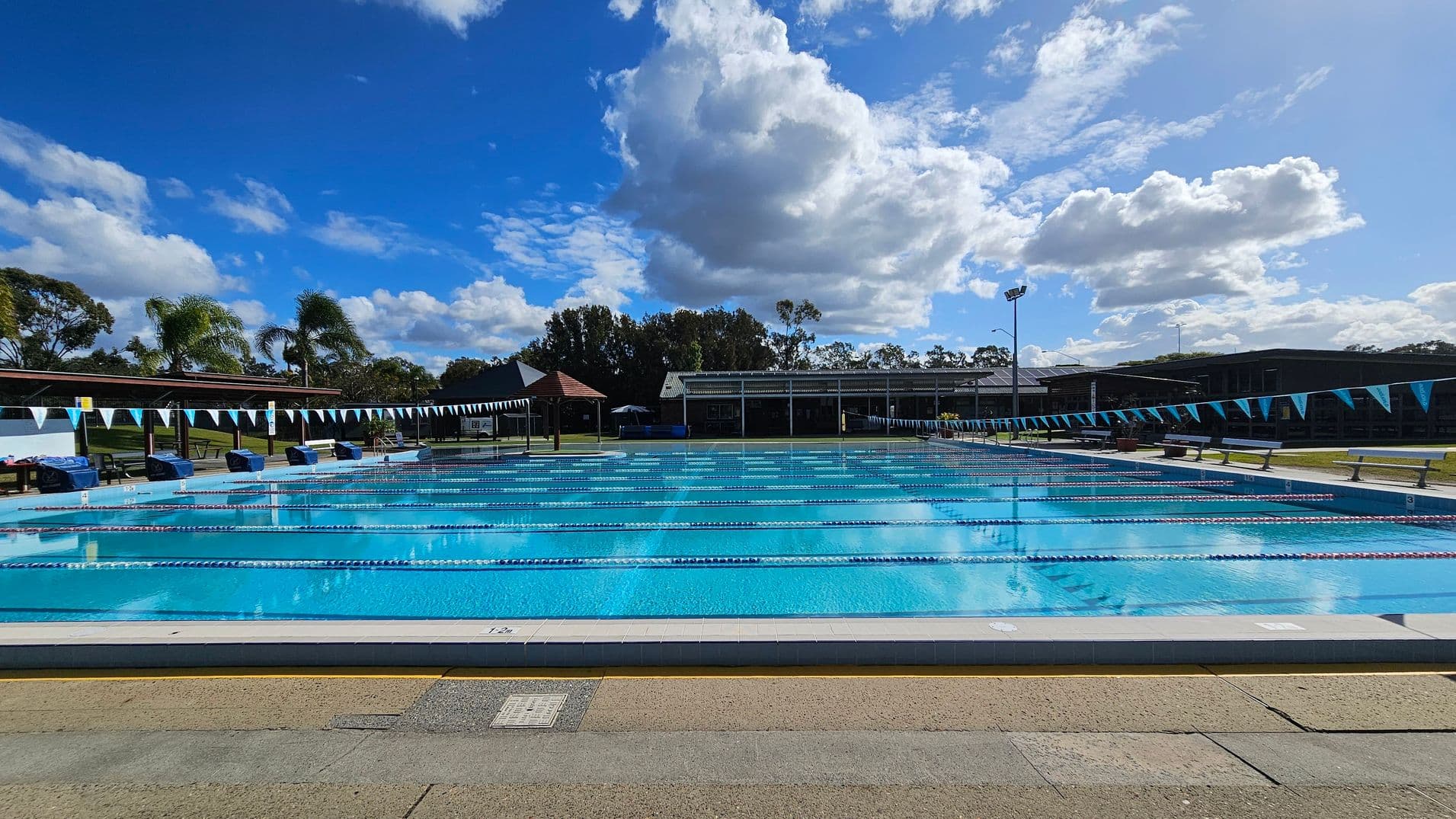 An outdoor swimming pool with clear blue water, divided into lanes by floating ropes. Blue and white triangular flags are strung across the pool. On the left and right, there are benches and a few trees, including palm trees. In the background, a building with a covered area and more trees is visible. The foreground shows a tiled pool deck with a mix of beige and green tiles.