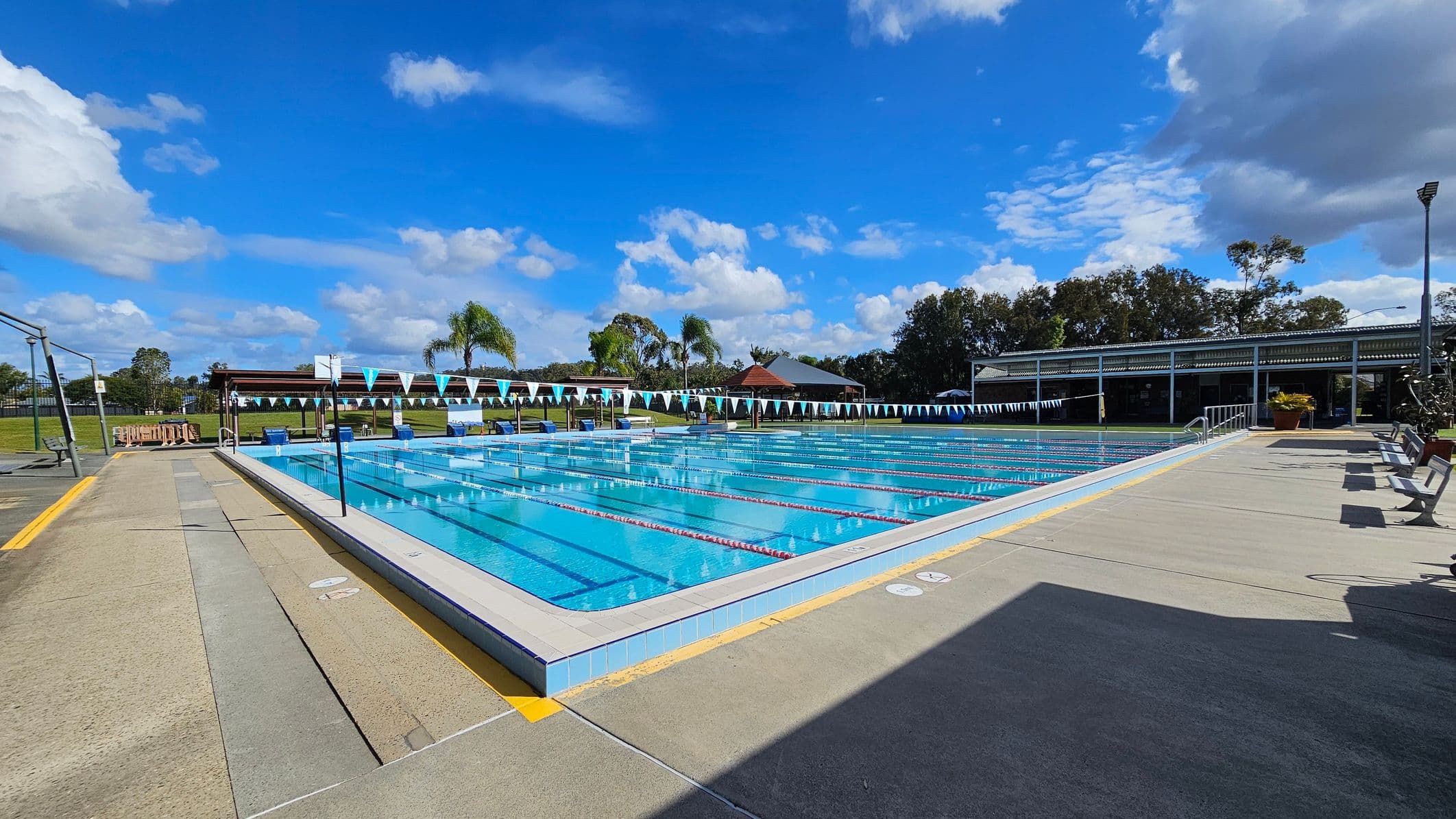 An outdoor swimming pool with multiple lanes, bordered by a wide concrete deck. On the left, there is a grassy area with a few palm trees and a wooden structure. On the right, a covered seating area with benches and potted plants. The pool is marked with lane dividers and overhead triangular flags. In the background, there are more trees and a building with a sloped roof. The sky is partly cloudy.