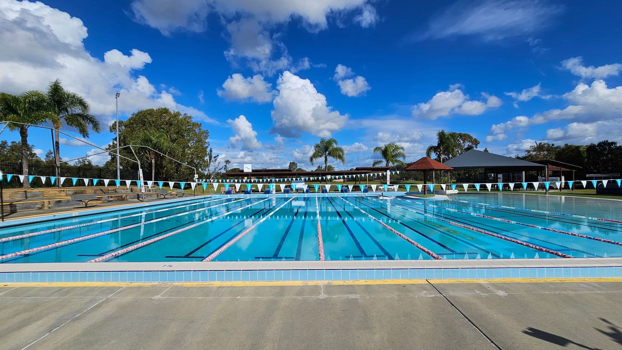 An outdoor swimming pool with multiple lanes, marked by blue and white flags strung across. The pool is surrounded by a concrete deck. In the background, there are palm trees and a few structures with red and grey roofs. Benches are visible along the left side, and the area is enclosed by a fence.