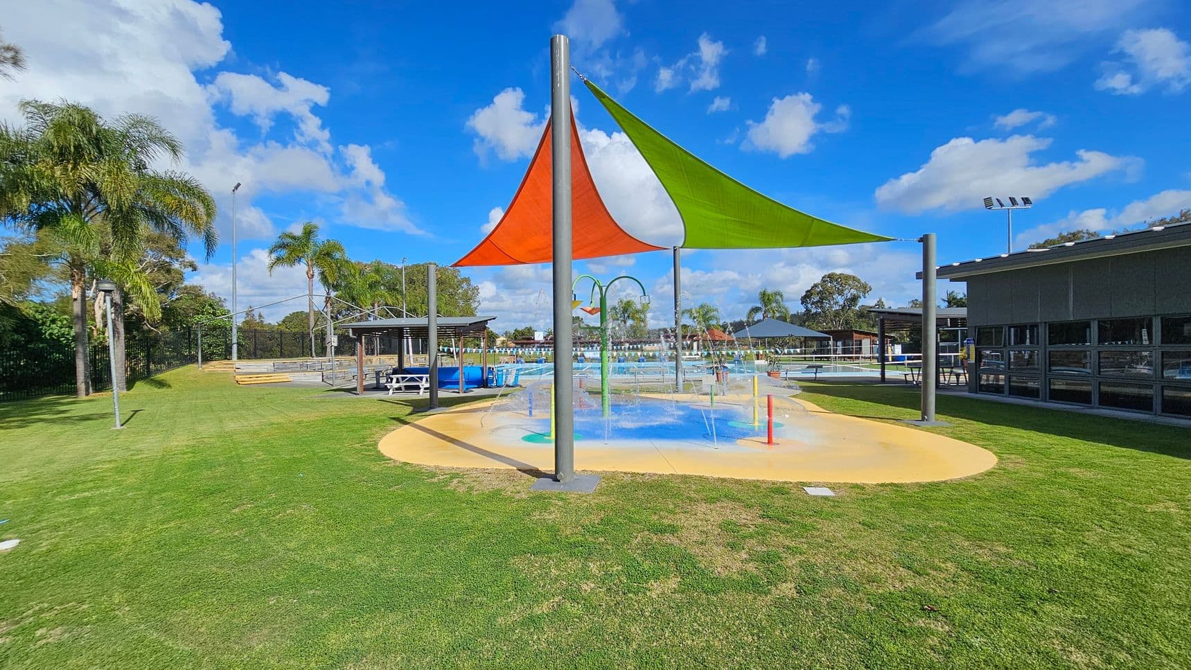 A splash pad with colourful triangular shade sails in orange and green. Water jets are spraying from the ground. To the left, there is a grassy area with palm trees and a few benches. In the background, a fenced area with more seating and a pool is visible. On the right, a building with large windows and a shaded seating area is present. The ground is a mix of grass and concrete, and the sky is partly cloudy.