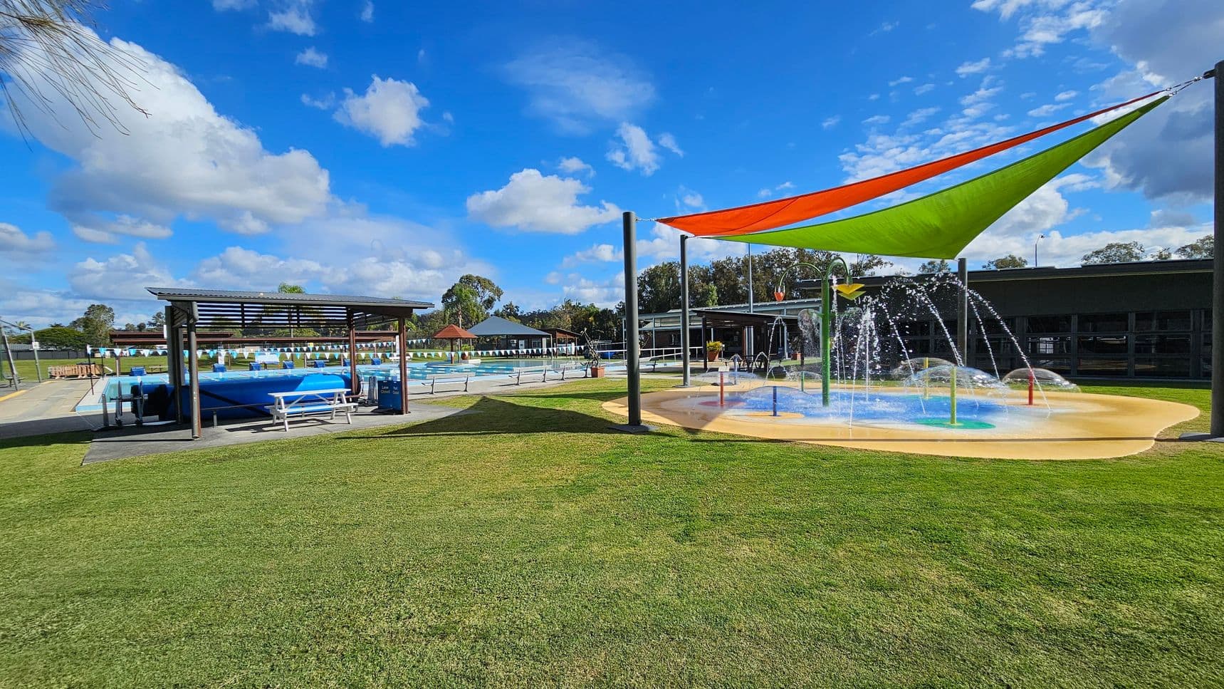On the left, there's a covered seating area with a picnic table and a blue pool cover nearby. In the centre, a large swimming pool is visible with lane dividers and a small building in the background. On the right, a splash pad with colourful water features is shaded by orange and green triangular canopies. The ground is covered with well-maintained grass, and there are trees and additional structures in the background.