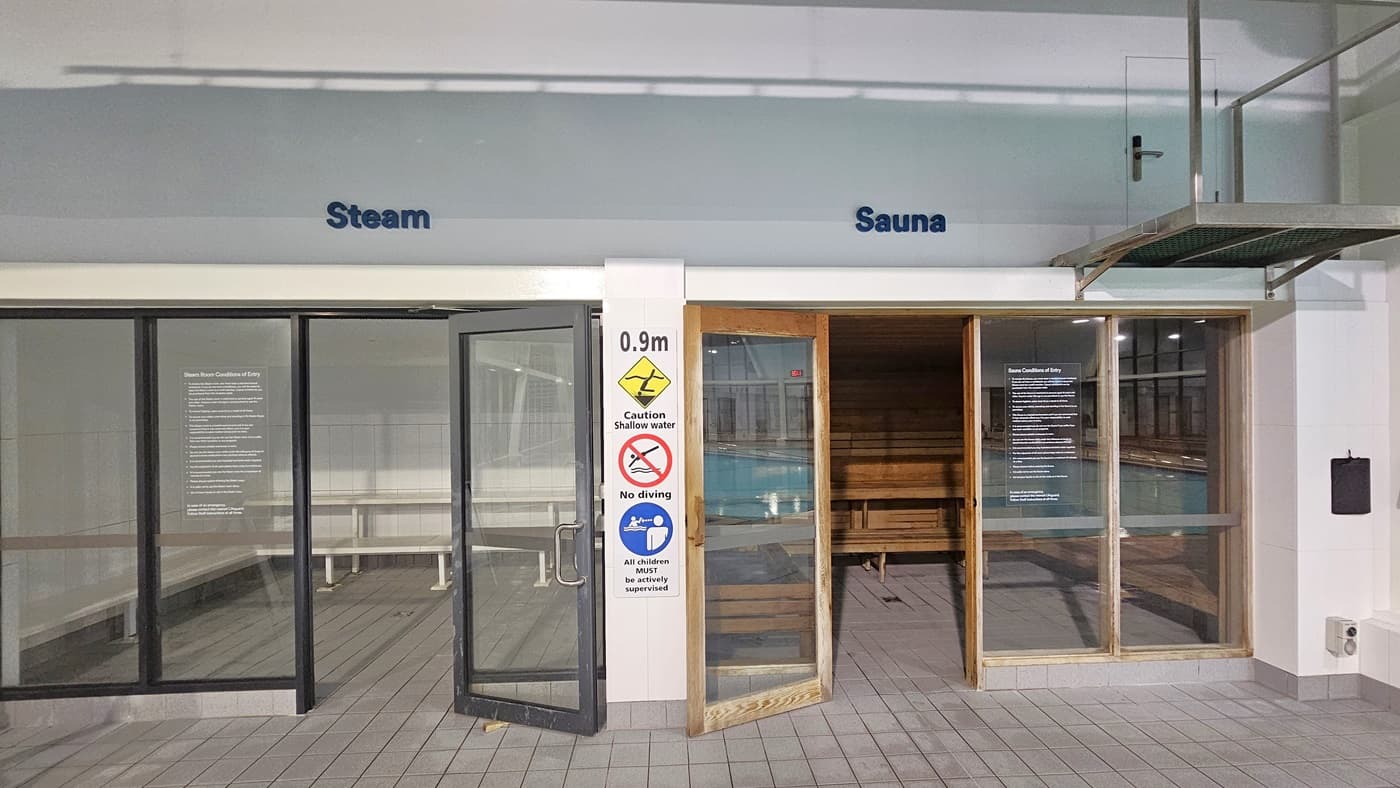 An interior view featuring two adjacent wellness facilities. On the left, a glass door with signage reading "Steam" leads to a steam room. A warning sign and a notice are affixed next to the entrance. To the right, an open wooden door with the word "Sauna" above indicates access to a sauna, showing wooden benches inside. In the foreground, a sign warns of a 0.9m deep no diving area. The ceiling hosts recessed lighting and a speaker, while the floor is tiled with grey.