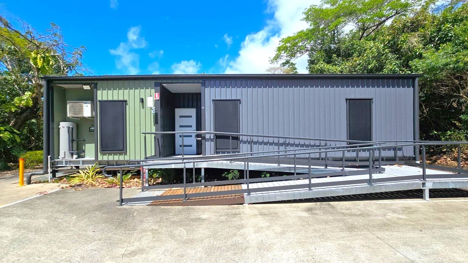 A small, rectangular building with a corrugated exterior, featuring a green section on the left and a grey section on the right. The building has a white door in the centre and two dark windows. A metal ramp with railings leads up to the entrance. An air conditioning unit is mounted on the left side. The foreground shows a concrete surface with scattered leaves, while the background is filled with dense green foliage and trees.
