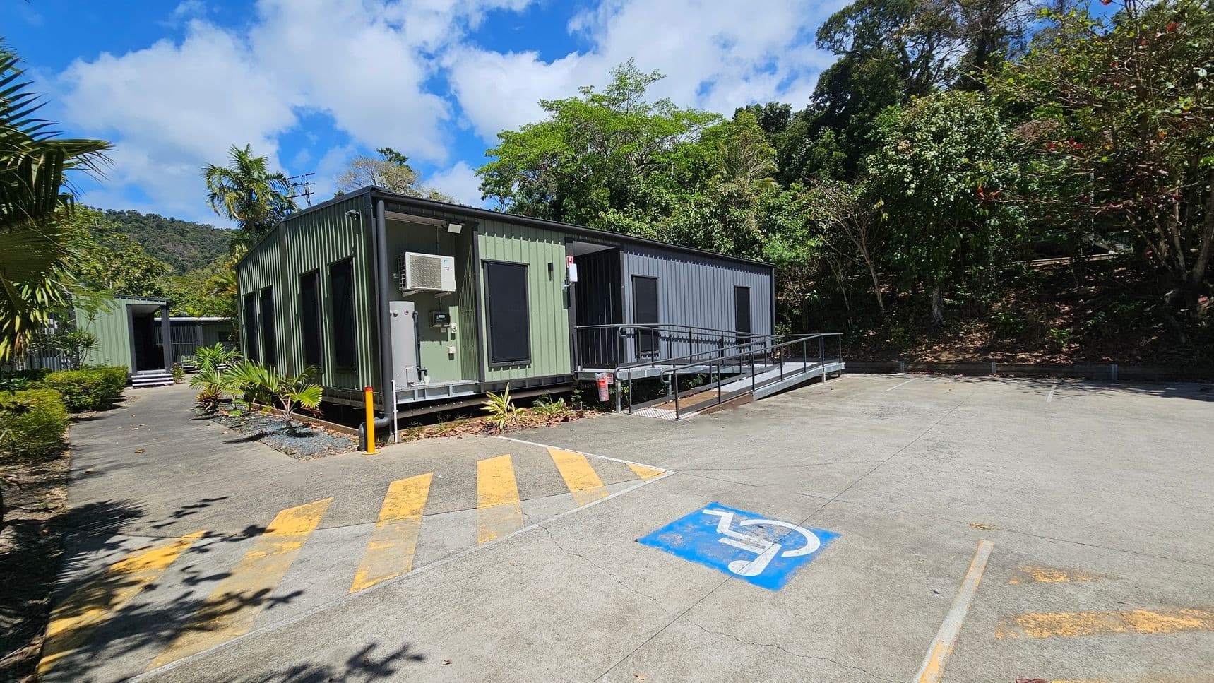 A green, rectangular building with large windows and a ramp is situated on the right. An air conditioning unit is mounted on the side. The foreground features a concrete area with a blue disabled parking symbol and yellow diagonal lines. On the left, a path leads to another building, surrounded by small plants and bushes. The background is filled with dense trees and foliage, with a hill visible in the distance.