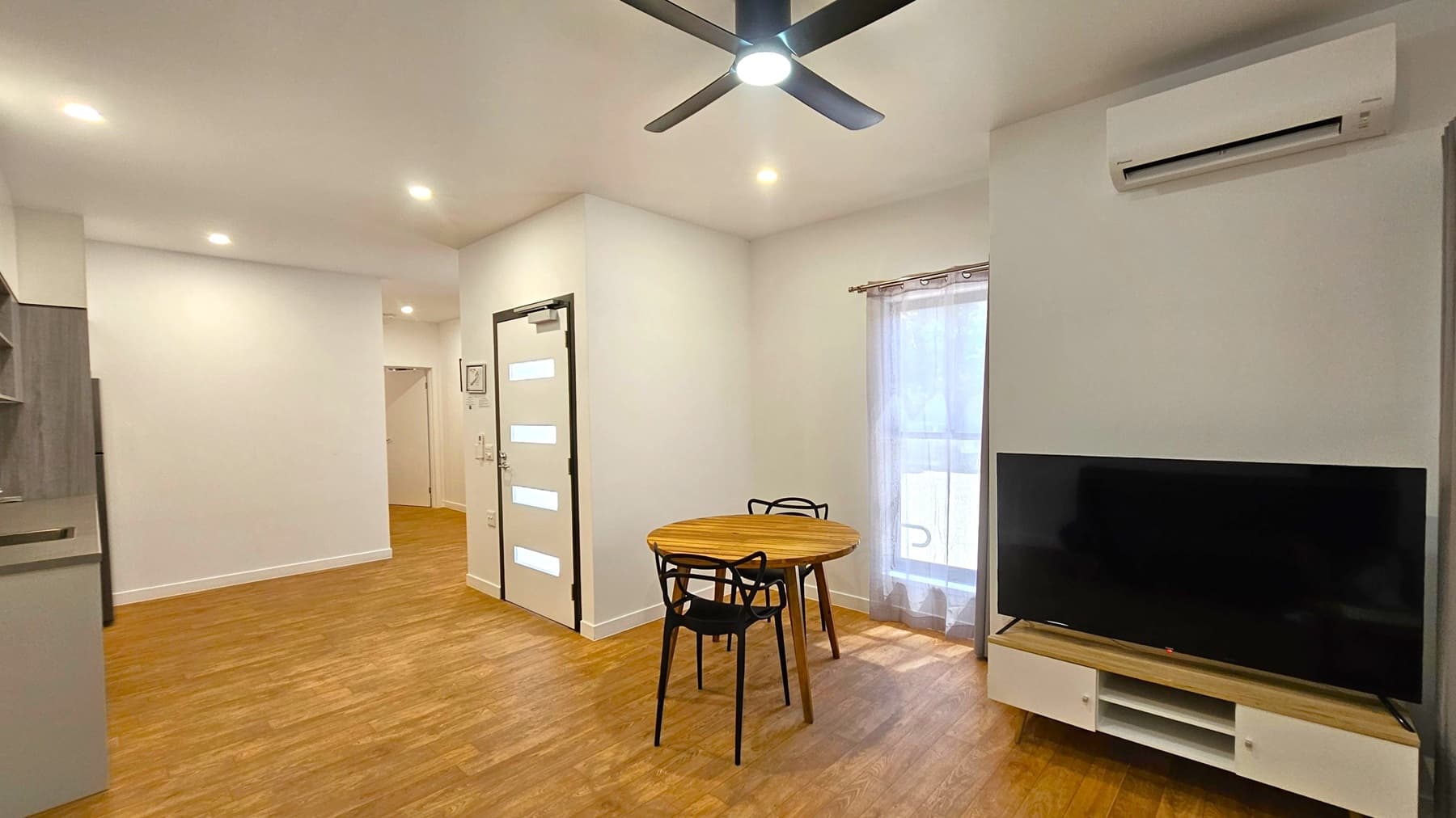 A modern living space with wooden flooring and a white ceiling featuring recessed lighting and a ceiling fan. On the right, there's a large flat-screen TV on a stand beneath an air conditioning unit. A round wooden table with two black chairs is positioned near a window with sheer curtains. On the left, part of a kitchen area is visible. A white door with horizontal glass panels is in the centre, leading to another room.