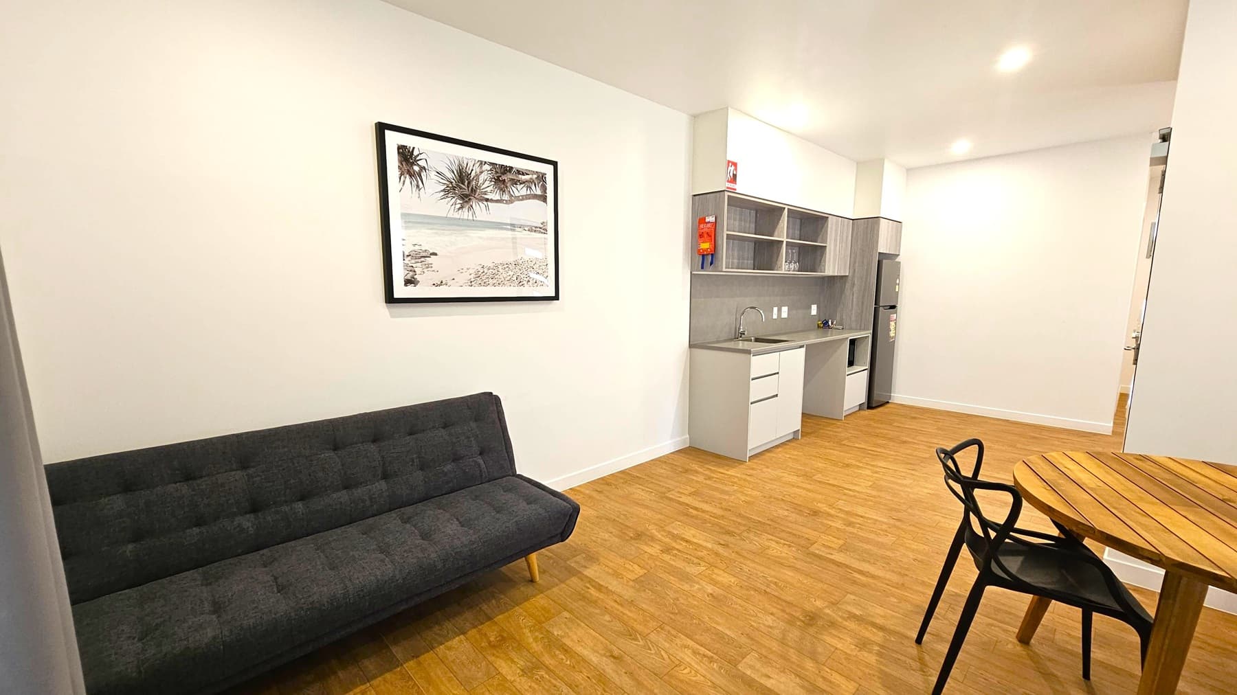 A room with light-coloured walls and a wooden floor. On the left, a dark grey sofa is positioned against the wall, above which hangs a framed beach photograph. In the centre, a compact kitchen area features light-coloured cabinets, a sink, and a small countertop. On the right, a round wooden table is accompanied by a black chair. The ceiling is white with recessed lighting.