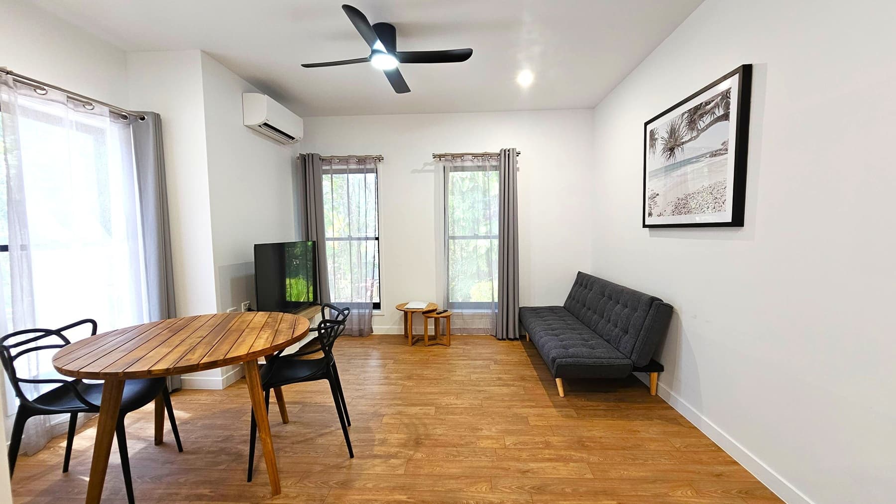 A modern living room with wooden flooring. On the left, a round wooden table with black chairs is near a window with sheer curtains. A TV is placed in the corner. In the centre, two tall windows with grey curtains allow natural light. On the right, a dark grey sofa sits against the wall, above which hangs a framed black-and-white beach photograph. A ceiling fan is mounted above, and a small air conditioning unit is on the wall.