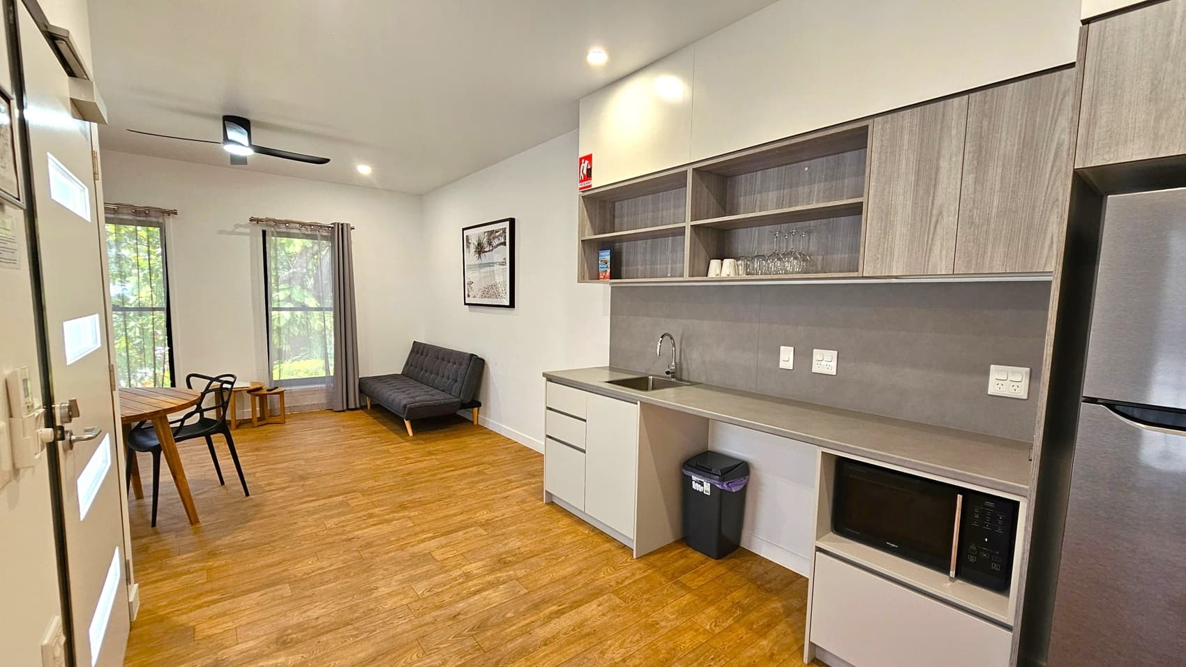 A modern room with wooden flooring and a light-coloured ceiling. On the right, there's a kitchenette with a sink, open shelves holding glasses and cups, and a microwave. A fridge is visible at the far right. In the centre, a black bin is placed under the counter. On the left, a small wooden dining table with black chairs is near two large windows with grey curtains. A black sofa sits against the wall, and a ceiling fan with a light is above. A framed picture hangs on the wall.