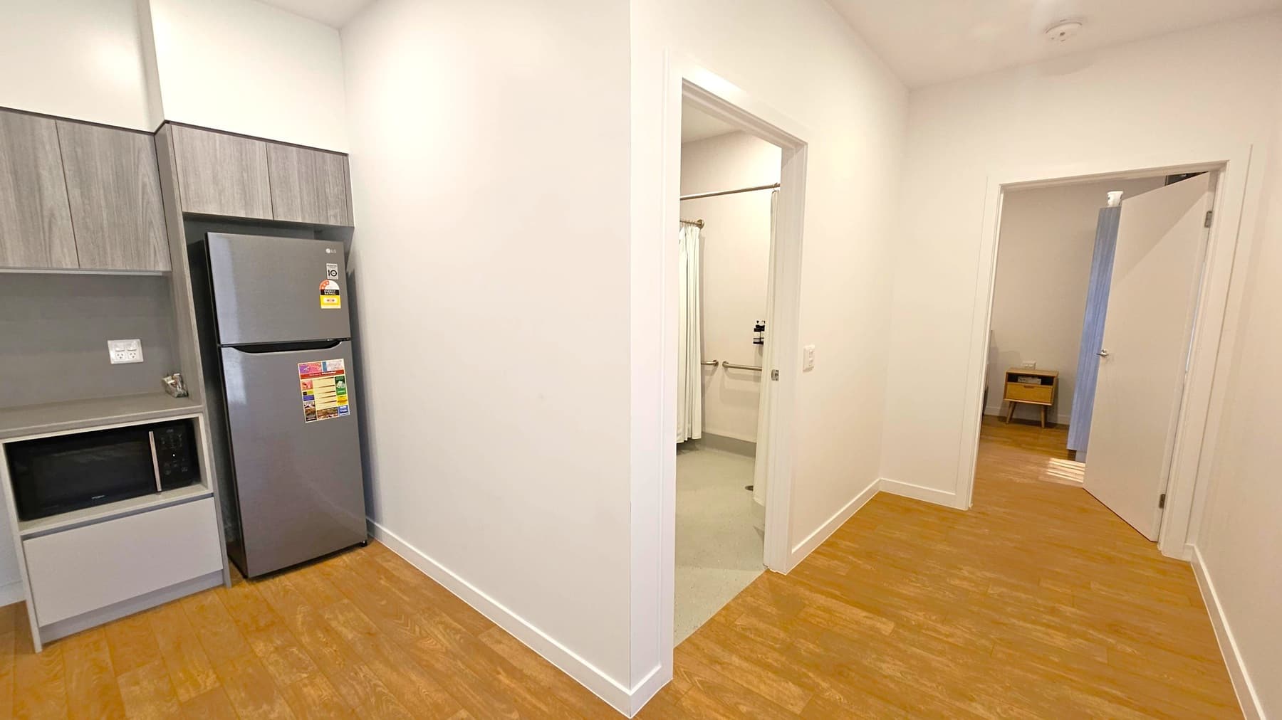 A small kitchen area on the left with a grey refrigerator, microwave, and wooden cabinets. The floor is wooden. In the centre, a hallway leads to a bathroom with a shower curtain. On the right, an open door reveals a room with a small table and a wooden floor. The walls are light-coloured, and the ceiling is plain with a smoke detector.